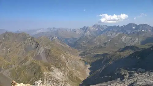 Vue sur une vallée et un lac de montagne depuis un sommet avec un ciel bleu et un nuage blanc au fond