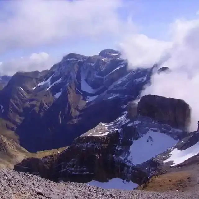 Vue panoramique sur les sommets enneigés du cirque de Gavarnie dans les Hautes-Pyrénées, avec un sentier de randonnée en premier plan et des nuages accrochés aux crêtes. Marmottes des Pyrénées .
