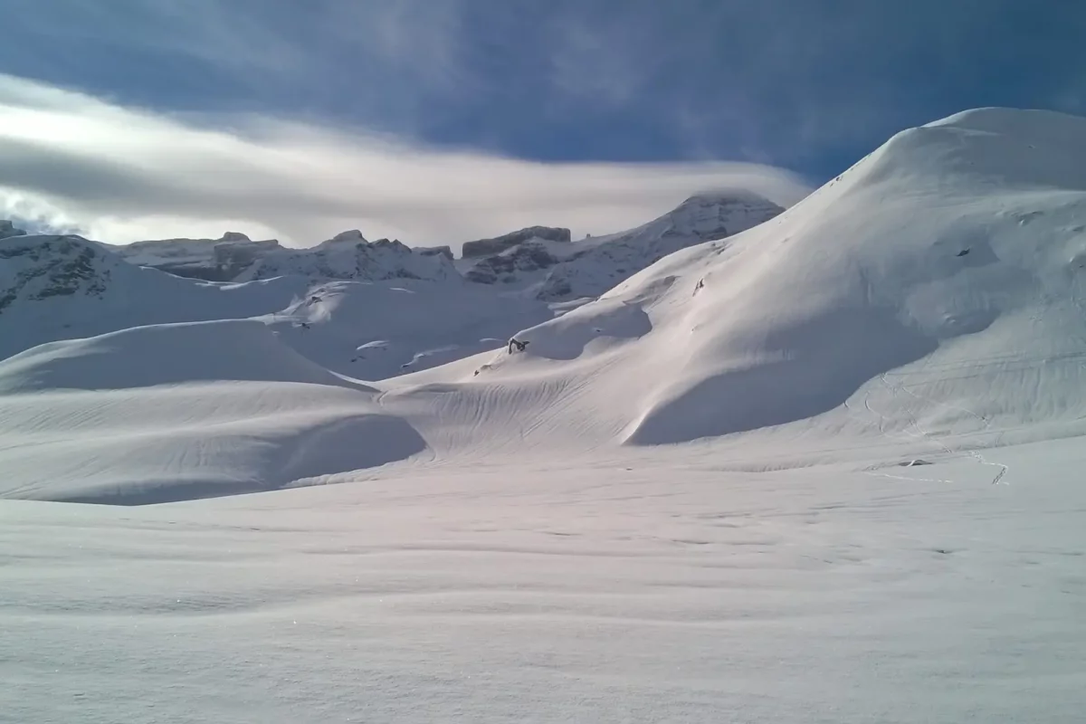 Vue sur la brèche de Roland depuis un paysage vallonée tout blanc avec un ciel bleu et les nuages qui accrochent les sommets du cirque de Gavarnie